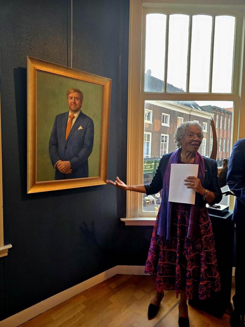 Foto van de Gouverneur van Curacao naast het nieuwe Staatsieportret, terwij zij haar speech houdt.