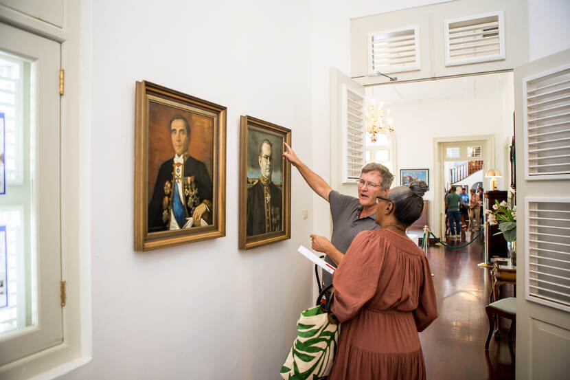 Een bezoeker van de openstelling is in gesprek met de directeur van het Kabinet. Zij staan in de portrettengalerij van het Paleis waar nu de portretten van Koloniale Gouverneurs hangen.